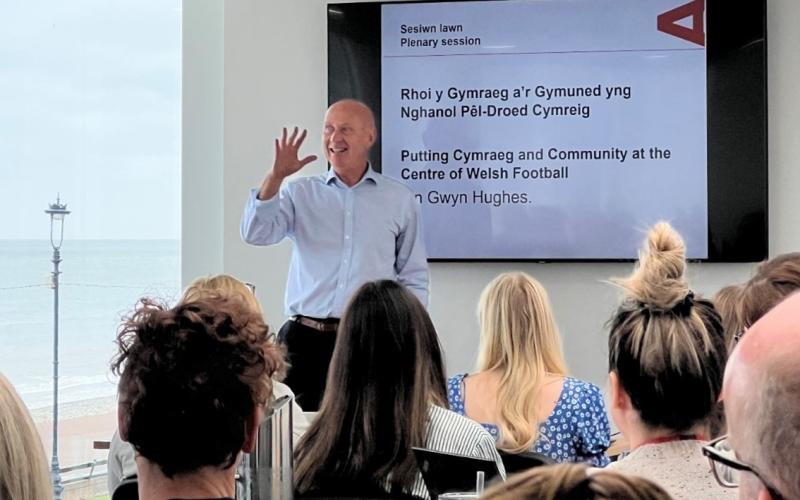 Ian Gwyn Hughes, Head of Communications at the FAW as the keynote speaker on stage talking. Text behind him saying Putting Cymraeg and Community at the centre of Welsh Football