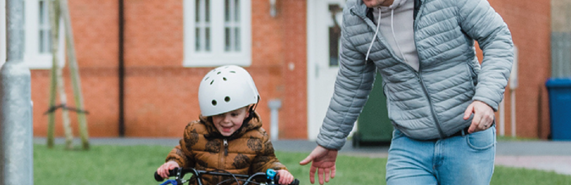 Picture of a father running with his young boy who is riding a bike with houses in the background.