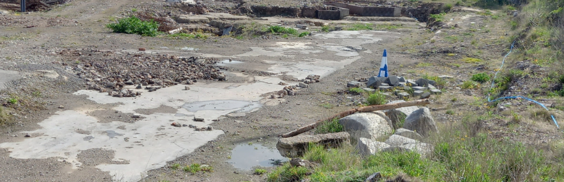 An empty, overgrown construction site with scattered grass and rocks.