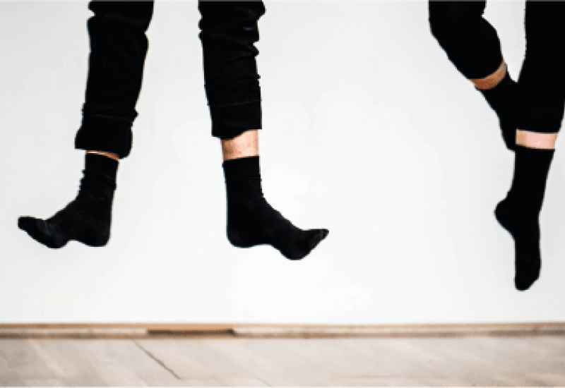 Photograph of two pairs of feet in black socks and black trousers, lifted from the ground and dancing